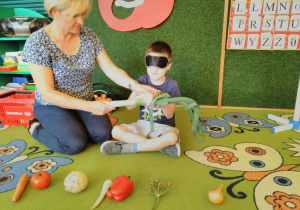 Dziecko rozpoznaje warzywo po dotyku, pod kierunkiem nauczycielki. Przed nim leżą warzywa.