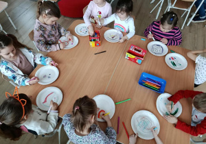 Dzieci siedzą przy stolikach, rysują wzory na papierowych talerzykach.