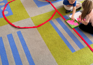 Dzieci siedzą na dywanie w grupach, układaja z obrazków cyfrę.