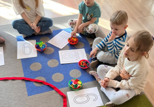 Dzieci siedża na dywanie z obrazkiem cyfry i miseczkami.