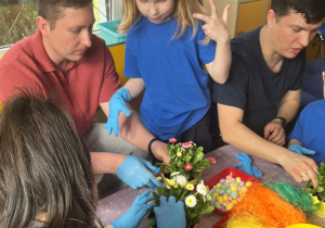 Rodzice z dziećmi wkładaja kwiat do doniczki.