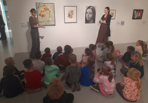 Dzieci siedzą na podłodze w muzeum i słuchają pracownic muzeum.