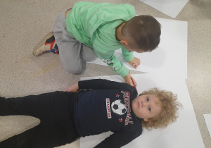 Chłopiec odrysowuje sylwetkę kolegi na białym papierze.