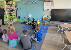 Dzieci siedzą na dywanie i słuchają strażnika miejskiego.