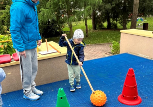 Chłopiec za pomoca kijka toczy piłkę.