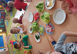 Dzieci siedzą przy stole i czekają na rozpoczęcie zajęć. Na stole są talerze z warzywami, chlebem, wędliną i serem, masło.