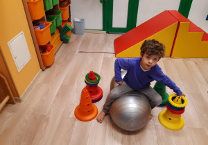 Chłopiec siedzi na piłce fasolce, balansując ciałem przekłada kółka z wypustkami sensorycznymi.