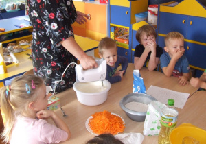 Nauczycielka wyrabia mikserem ciasto. Dzieci z ciekawością obserwują.