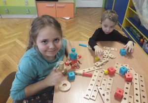 Dzieci siedzą przy stoliku i dziewczynka pokazuje swoją budowlę