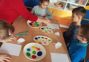 Dzieci przy stole na białej kartce odbijają paluszki zamoczone z farbą robiąc kropki.