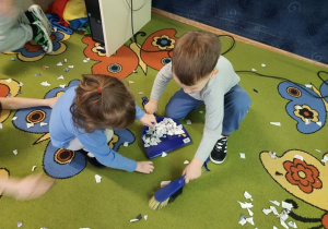 Dzieci zamiatają papierki z dywanu uzywając szczotki i szufelki.