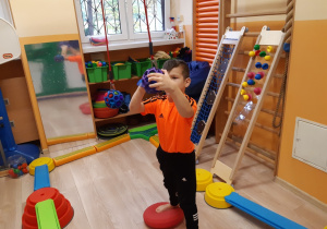 Chłopiec na dyskach sensorycznych wkłada kolorową chustkę w podwieszaną piłkę z otworami.