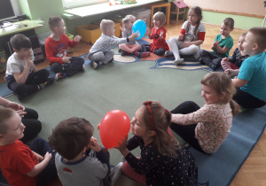 Dzieci przekazują sobie balon z rąk do rąk siedząc na dywanie.