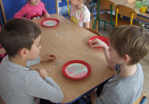 Dzieci siedzą przy stoliku, na stole leżą talerze z mlekiem.