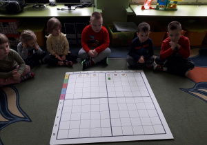 Dzieci siedzą na dywanie a przed nimi leży plansza do kodowania.