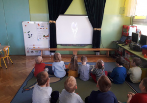 Dzieci siedzą na dywanie i oglądają prezentację multimedialną.