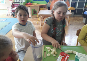 Dziewczynka kroi banana a chłopiec wkłada pokrojone kawałki do blendera.