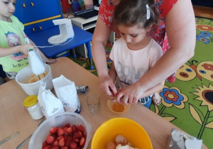 Nauczycielka pomaga dziewczynce rozbić jajko do miski.