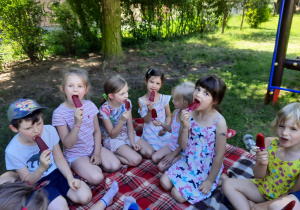 Dzieci siedzą na kocu. Dzieci jedzą lody.