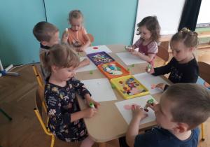 Dzieci siedzą przy stoliku i przyklejają kolorowe kółka na kartkę papieru.