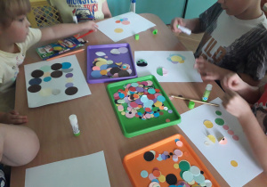 Dzieci siedzą przy stoliku i przyklejają kolorowe kółka na kartkę papieru.