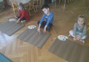 Dzieci siedzą na podłodze przed kartką szarego papieru.