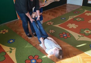 Nauczyciel przytrzymuje dziecko za nogi na wysokości kostek, lekko przeciąga je w różnych kierunkach.