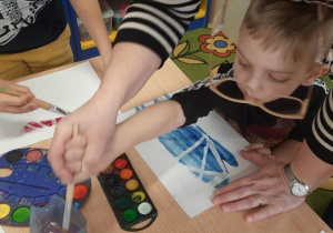 Chłopiec z pomocą nauczycielki maluje po kartce.