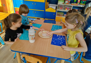 Dzieci palcami maczanymi w białej farbie robia kropki na niebieskiej kartce.