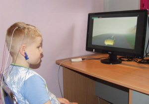 Dziecko siedzi przed monitorem, ma podłączone elektrody do głowy. Patrzy na zmieniający się obraz