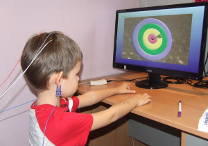 Dziecko siedzi przed monitorem, ma podłączone elektrody do głowy. Patrzy na zmieniający się obraz